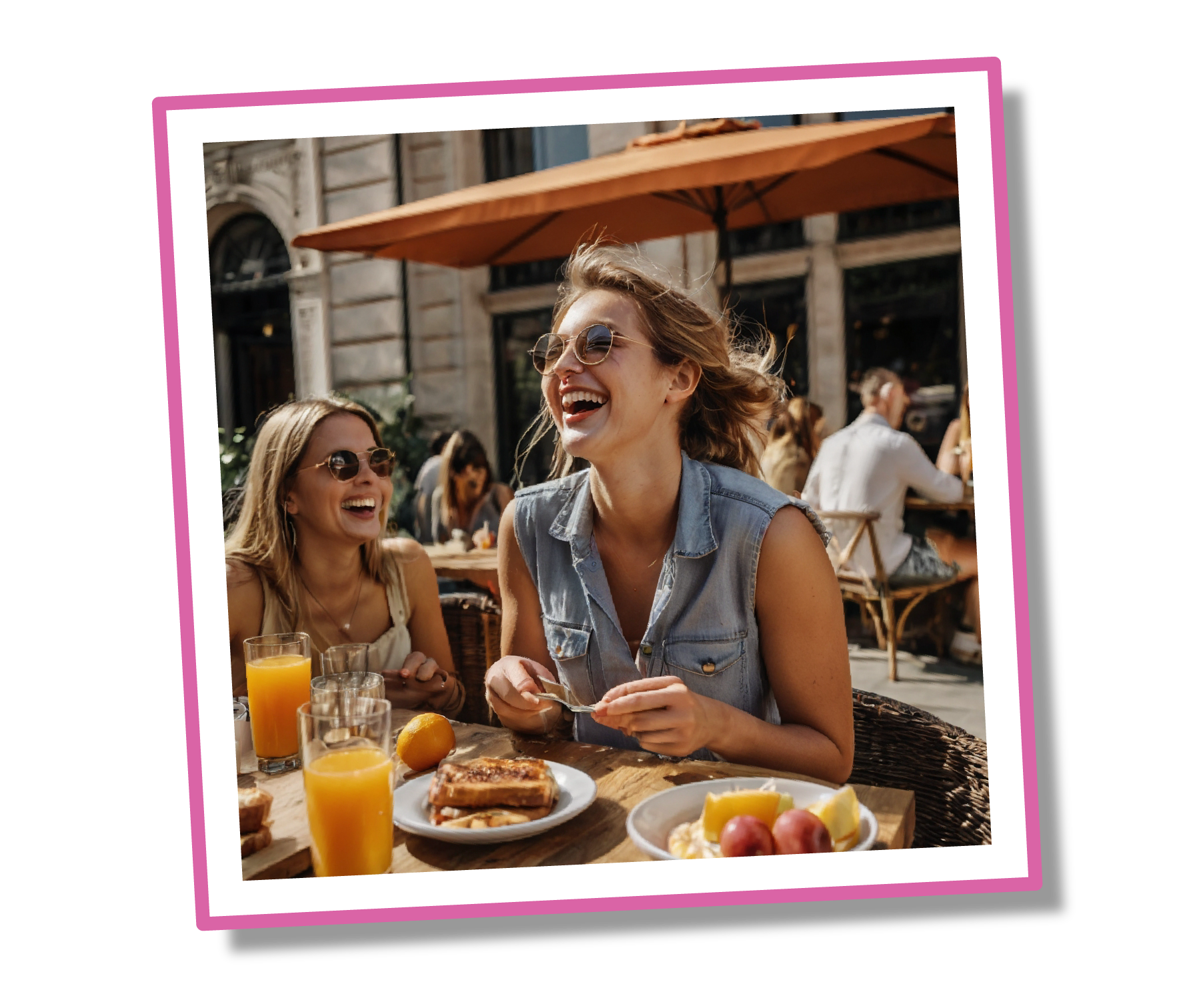 Two women laughing at an outdoor cafe table with food and drinks.