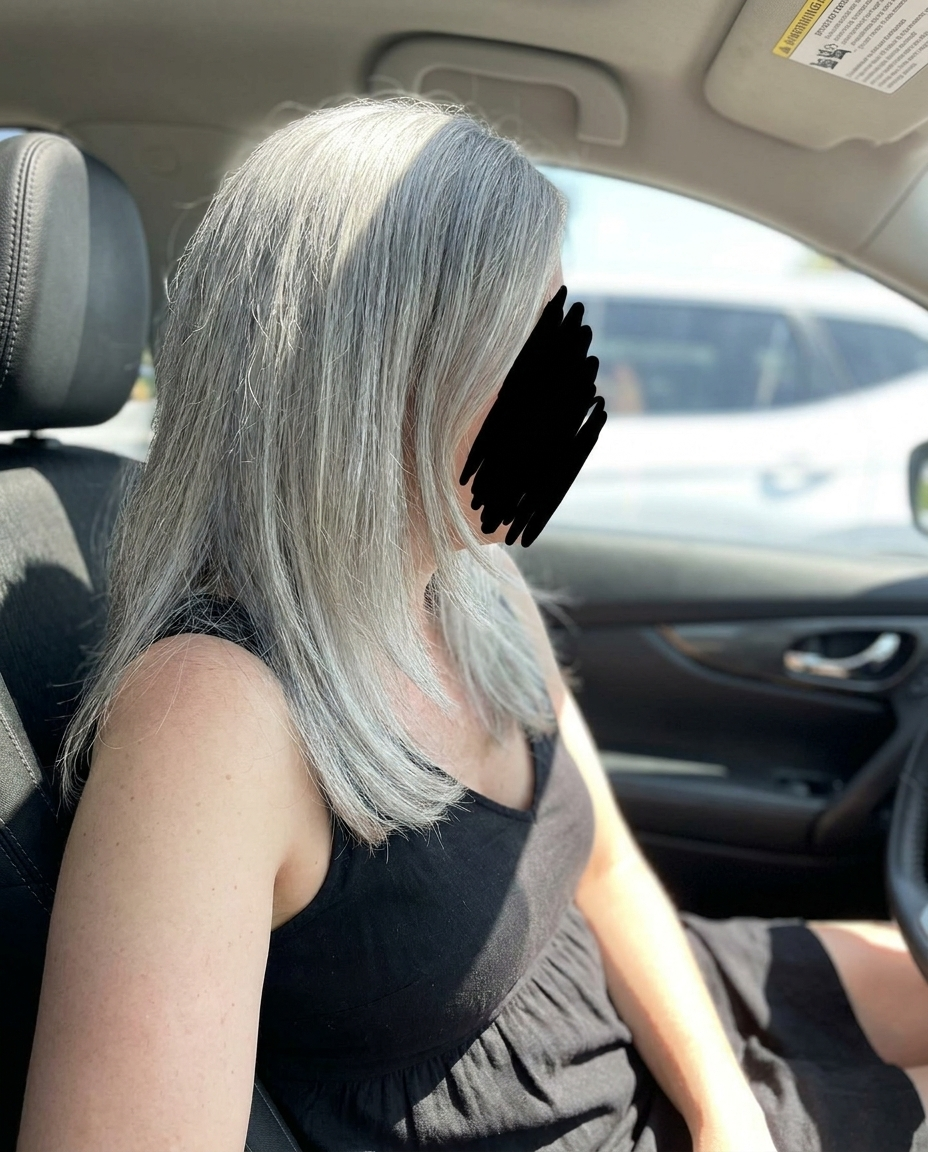 A person with long gray hair, wearing a black top, sits in the driver's seat of a car.