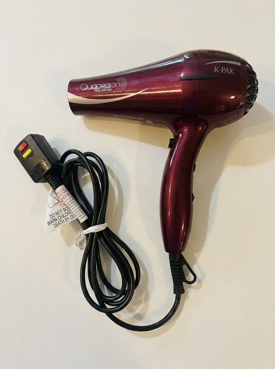 A maroon K-PAK Quadraion Pro-Dryer hair dryer with its cord bundled, lying on a white surface.