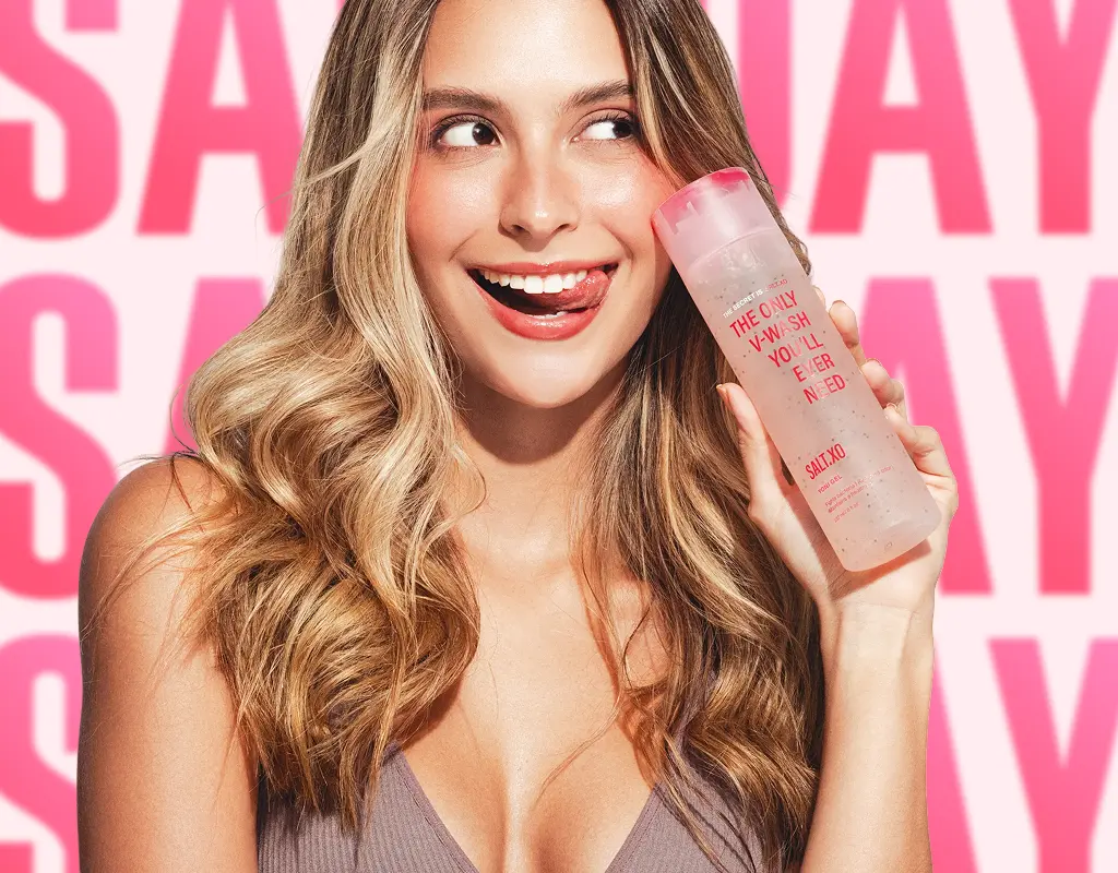 A smiling woman with blonde hair holds a bottle of SALT.XO V-Wash against a pink background.