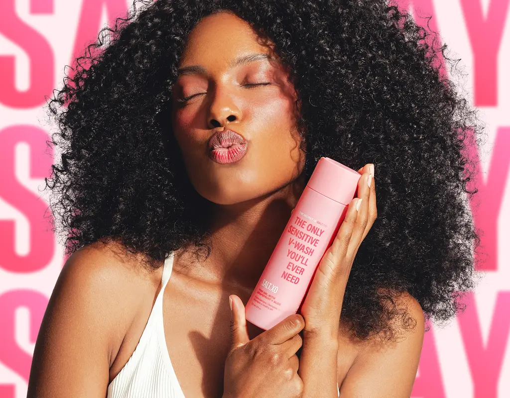 A woman with curly hair puckers her lips while holding a pink bottle of 'The Only Sensitive V-Wash'.
