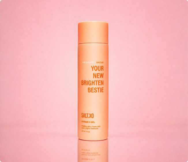 An orange bottle of Salt.XO Vitamin C Gel against a solid pink background with its reflection showing below.