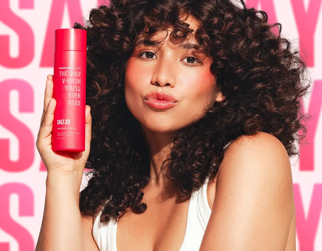 A woman with curly hair holds a pink bottle of Salt.xo v-wash against a pink background.
