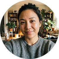 A woman in a grey sweater smiles in a room filled with bookshelves and plants.