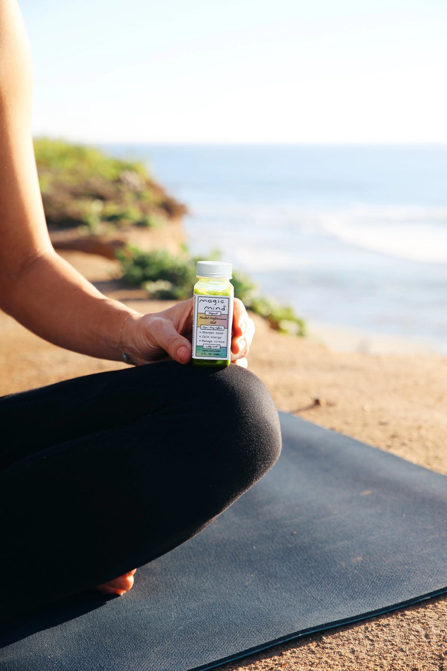 Person holding a small bottle labeled 'Magic Mind' on a yoga mat by the beach.