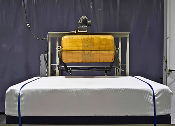 Machine testing a mattress with a wooden roller in a laboratory setting.