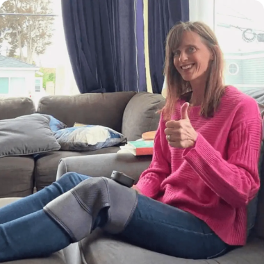 A smiling woman sits on a couch wearing a knee brace and giving a thumbs-up.