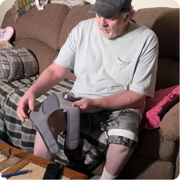An older man sitting on a couch holds and looks down at a therapeutic knee brace.