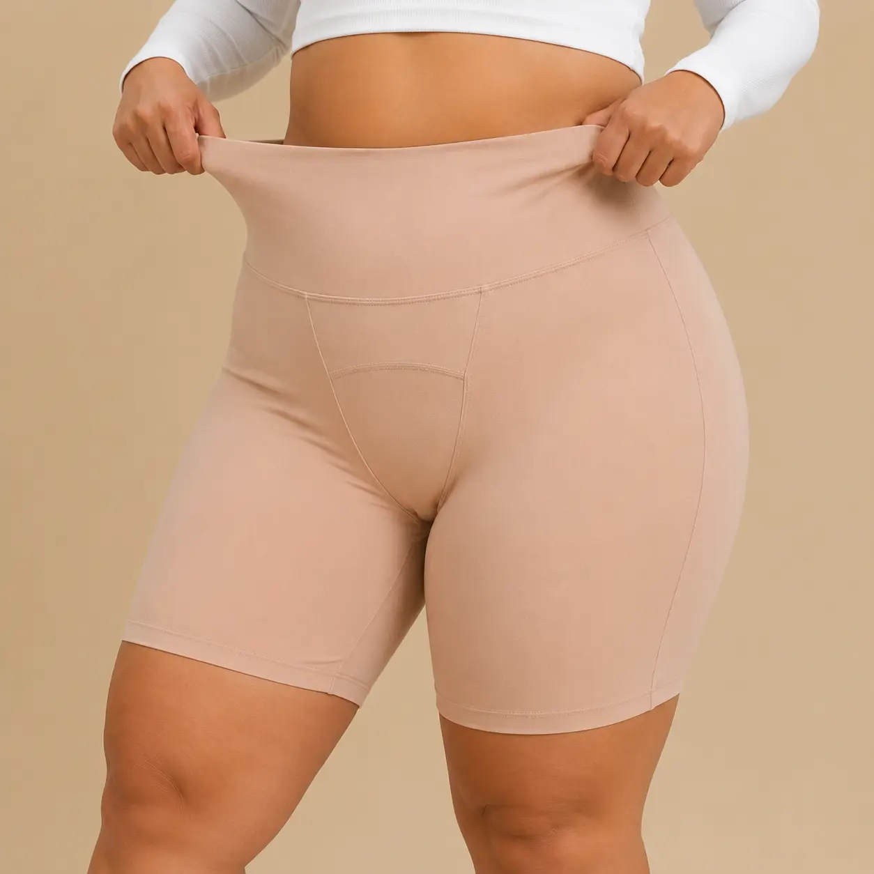 A woman in a white top stretches the waistband of her light brown, high-waisted biker shorts.
