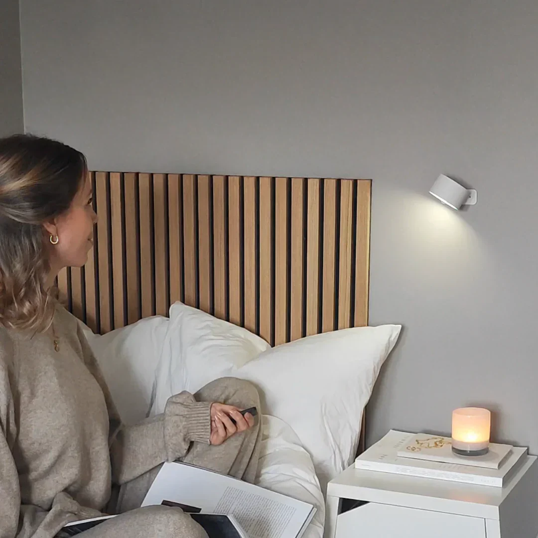 Person sitting on bed with book, beside a lit candle and wall light.