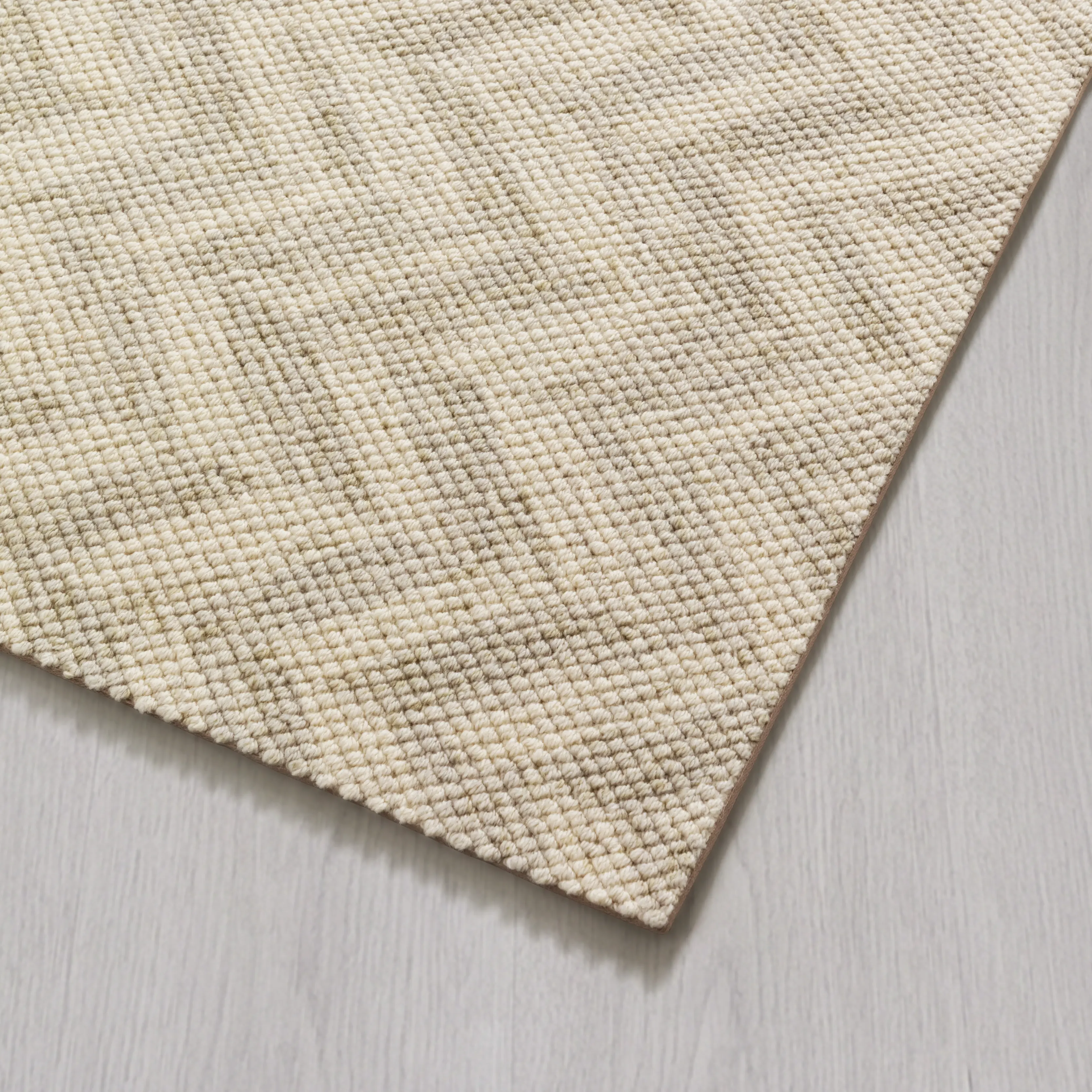 A close-up of the corner of a textured, beige and gray woven rug on a light wood floor.