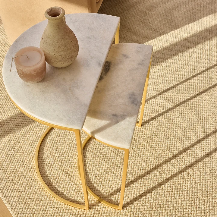 Two semi-circular nesting tables with white marble tops and gold frames, holding a vase and candle.