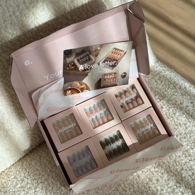 An open pink box filled with six smaller packages of Glowie brand press-on nails on a blanket.