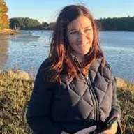 A smiling woman with reddish-brown hair in a black quilted vest, standing outdoors by a body of water.