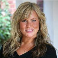 A headshot of a smiling woman with wavy blonde hair and a dark V-neck shirt.
