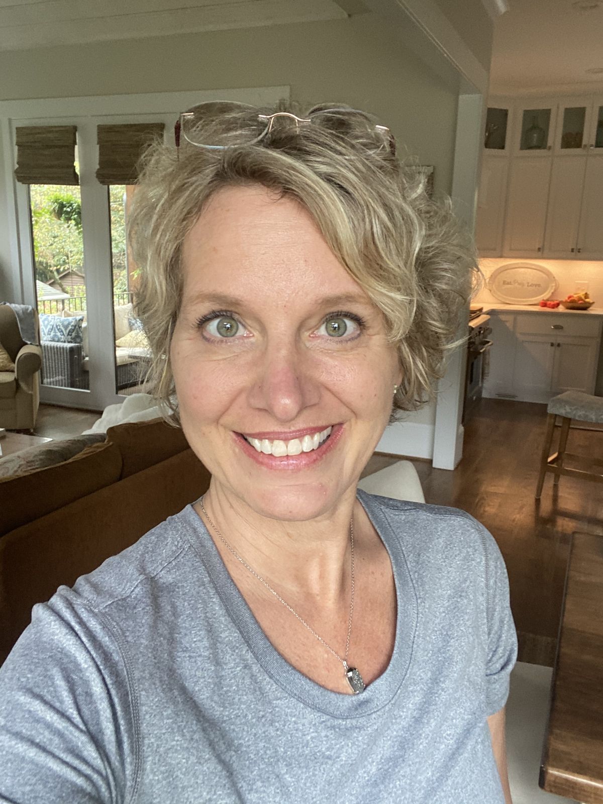 A smiling woman with short, curly blonde hair takes a selfie indoors in her living room.