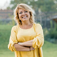 A smiling blonde woman in a yellow shirt stands outside with her arms crossed.