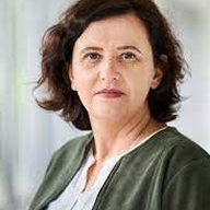 A headshot of a woman with dark wavy hair, wearing a green jacket and a white blouse.