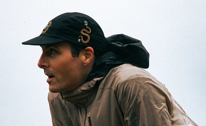 A man in profile wearing a black embroidered cap and a tan hooded jacket against a light sky.