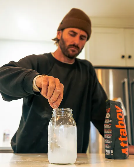 A man in a beanie sprinkles a substance into a glass jar on a kitchen counter.