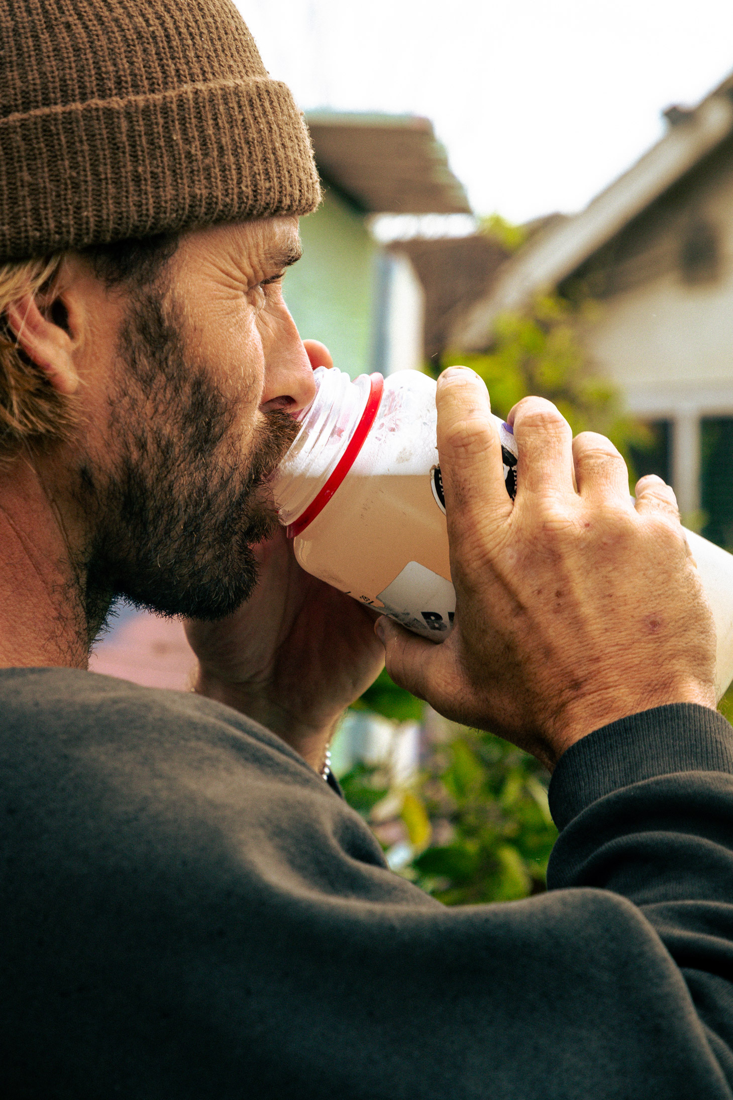 A man in a brown beanie and dark sweatshirt drinks from a plastic bottle outdoors.