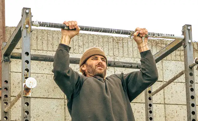 A man with a beard and beanie hangs from a pull-up bar on an outdoor rack.