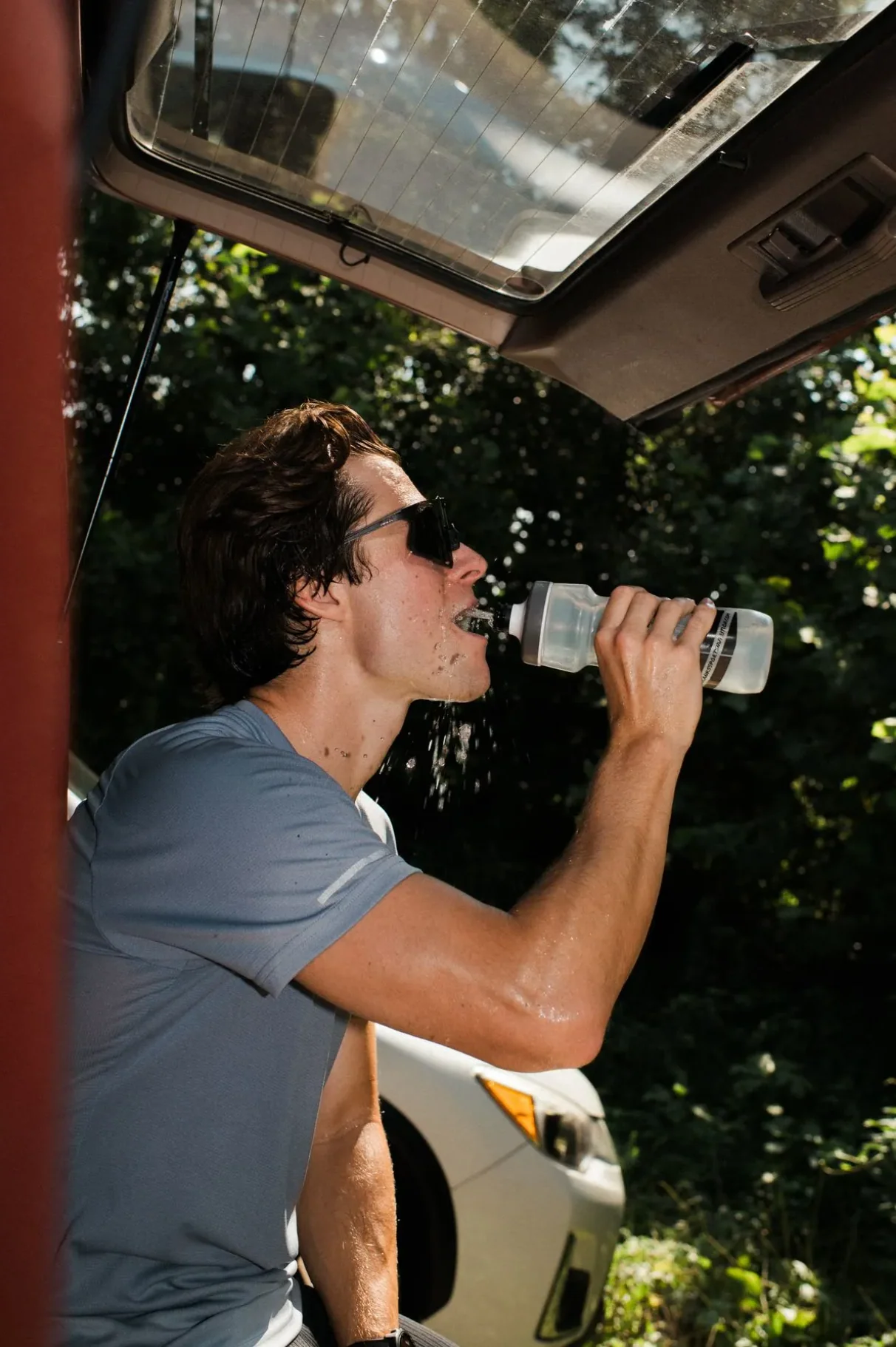 Person in sunglasses drinking water from a bottle under an open car trunk.
