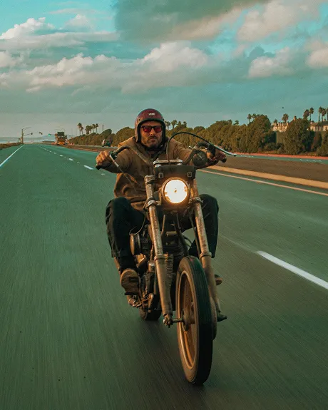 A man wearing a helmet and sunglasses rides a chopper motorcycle down an open, tree-lined road.