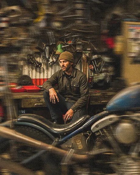 A man in a beanie sits on a motorcycle in a cluttered, blurry workshop.