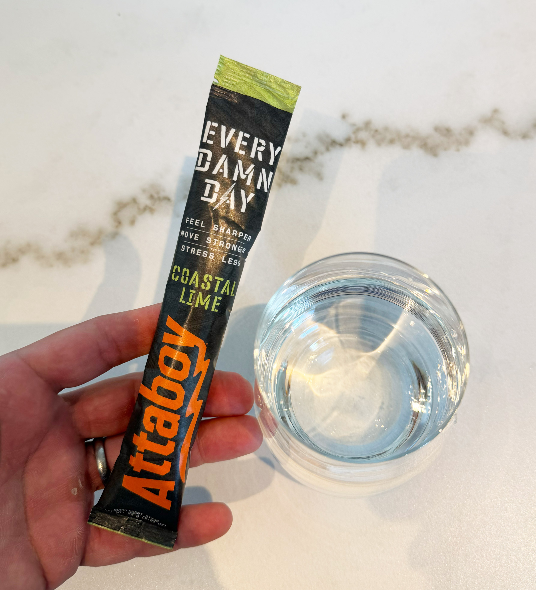 Hand holding a black packet labeled 'Every Damn Day,' beside a glass of clear liquid.