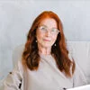 Person with long red hair sitting in a light-colored room.