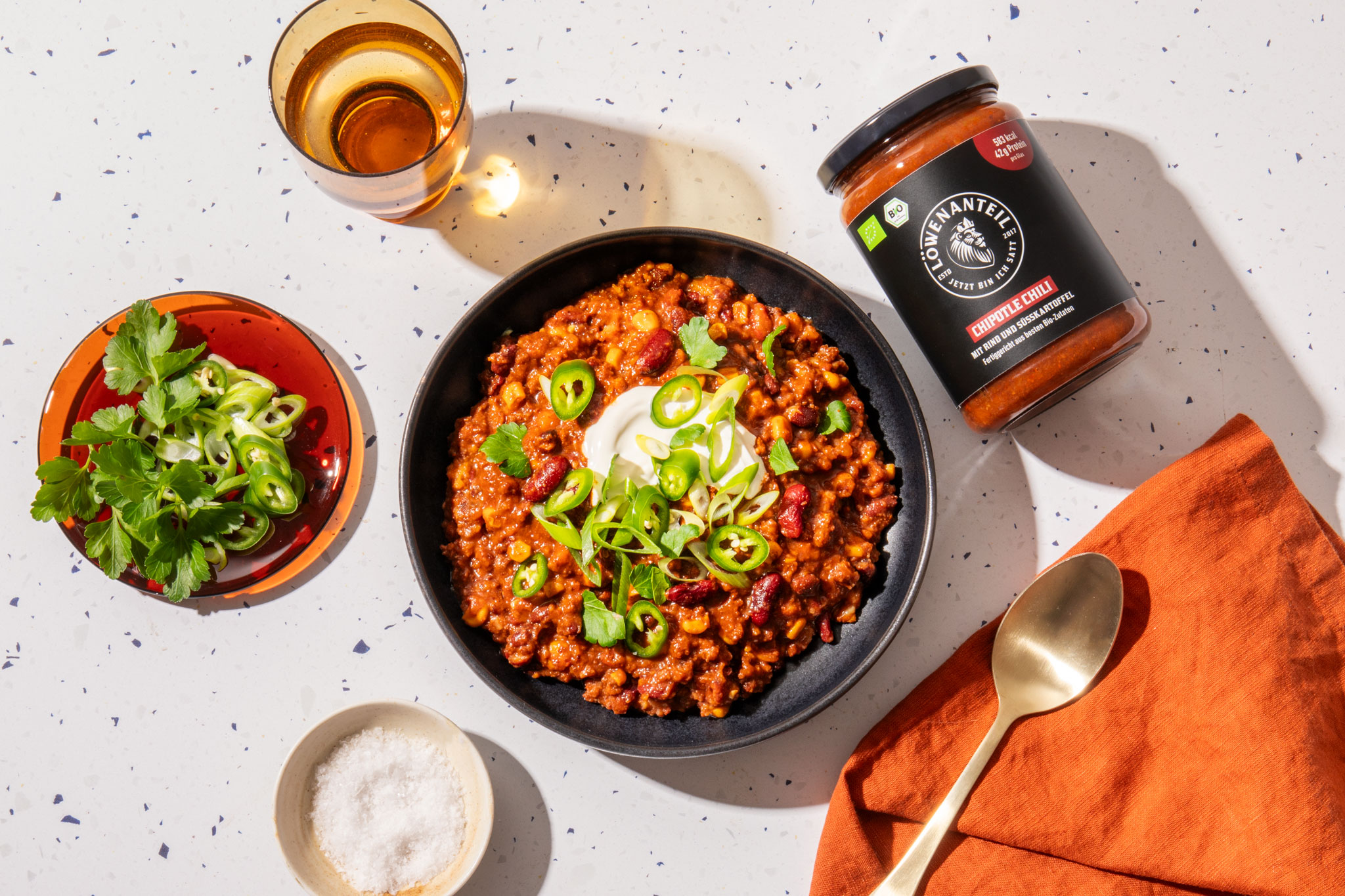 A bowl of chili topped with sour cream and jalapeños, next to a jar of chipotle chili sauce.