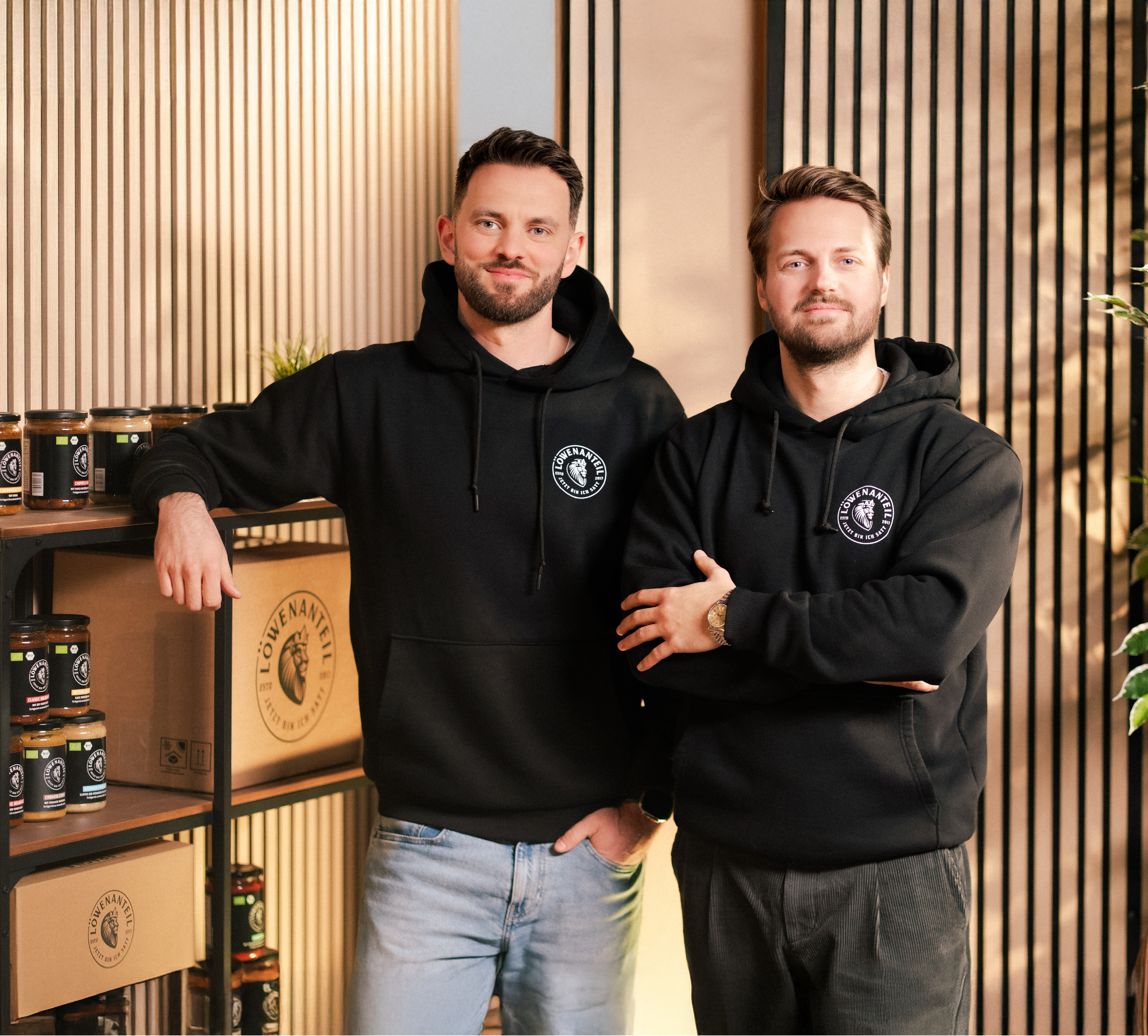 Two men in black hoodies standing, with shelves of jars and boxes in the background.
