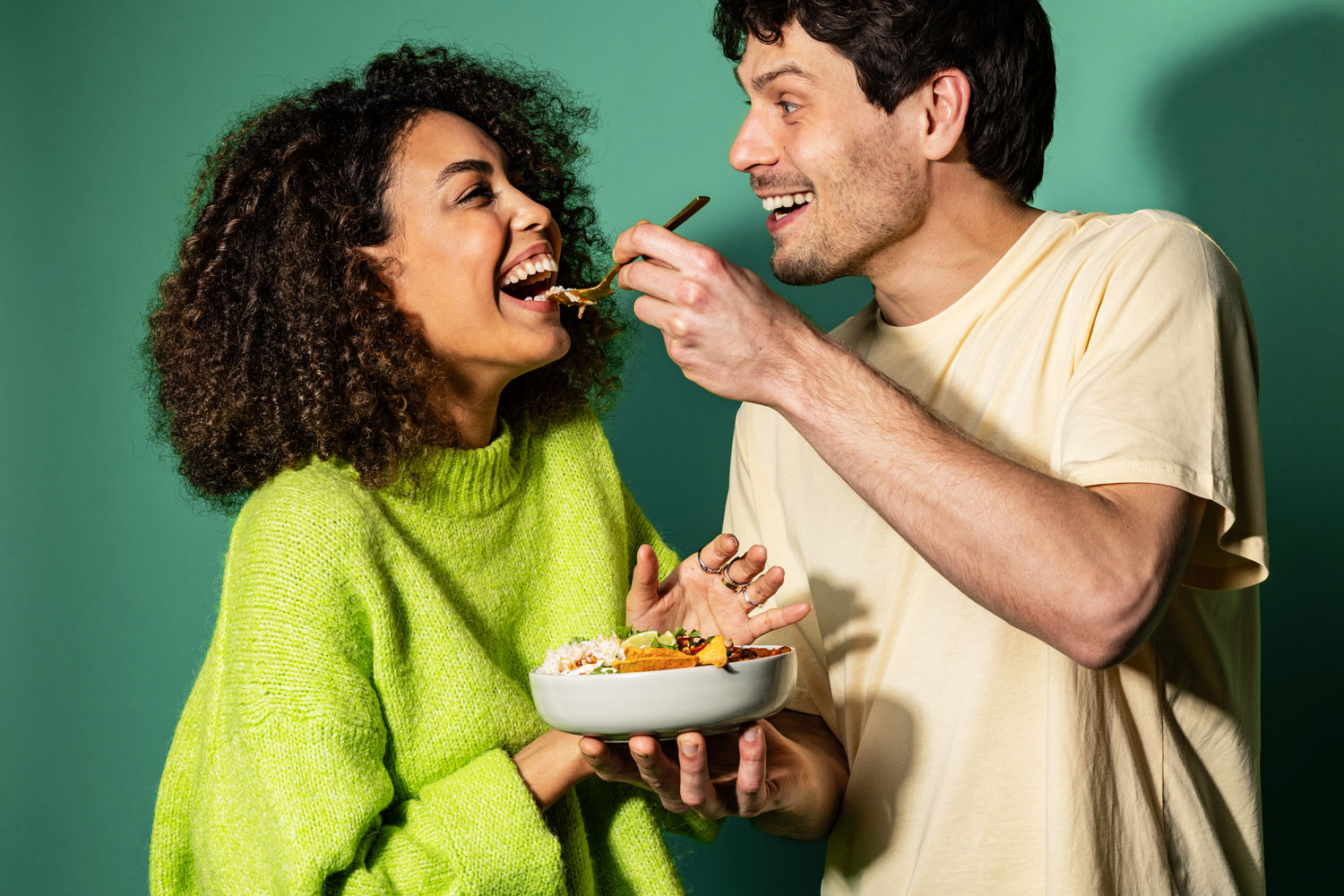 Two people laughing, one feeding the other with a spoon, holding a bowl of food.