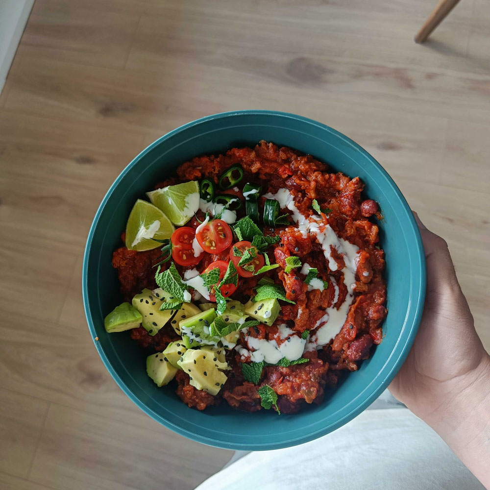 A person holding a teal bowl of chili topped with avocado, tomatoes, lime, and mint.