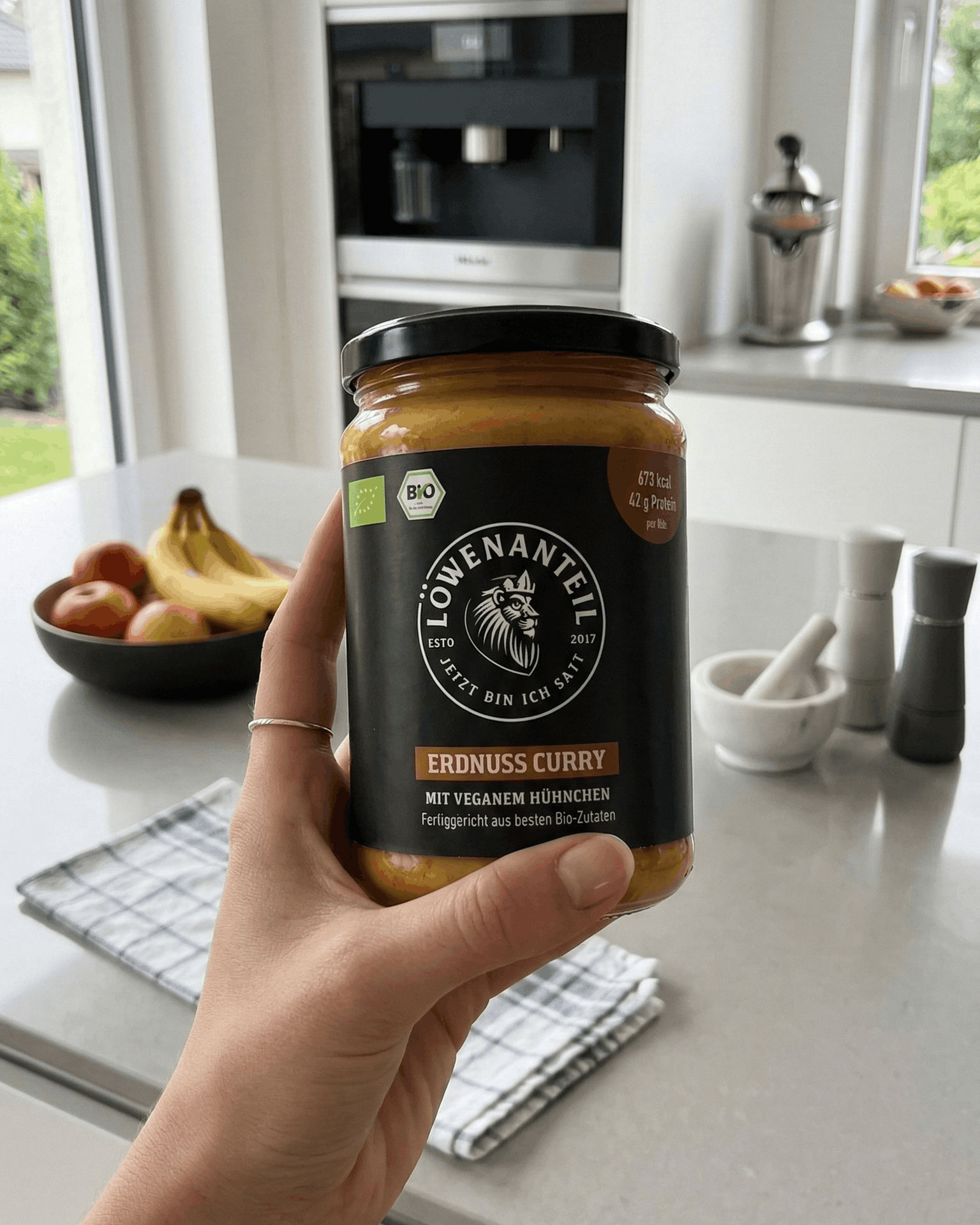 A person's hand holding a jar of Löwenanteil Erdnuss Curry (peanut curry) in a kitchen.