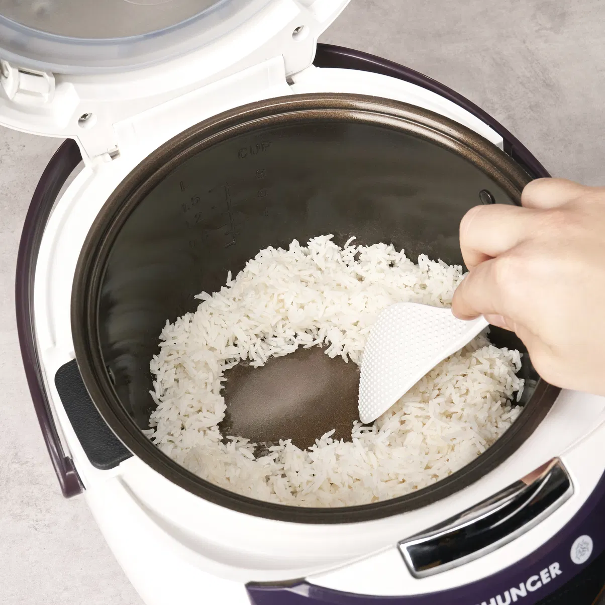 A hand scoops freshly cooked white rice from an open rice cooker with a white paddle.