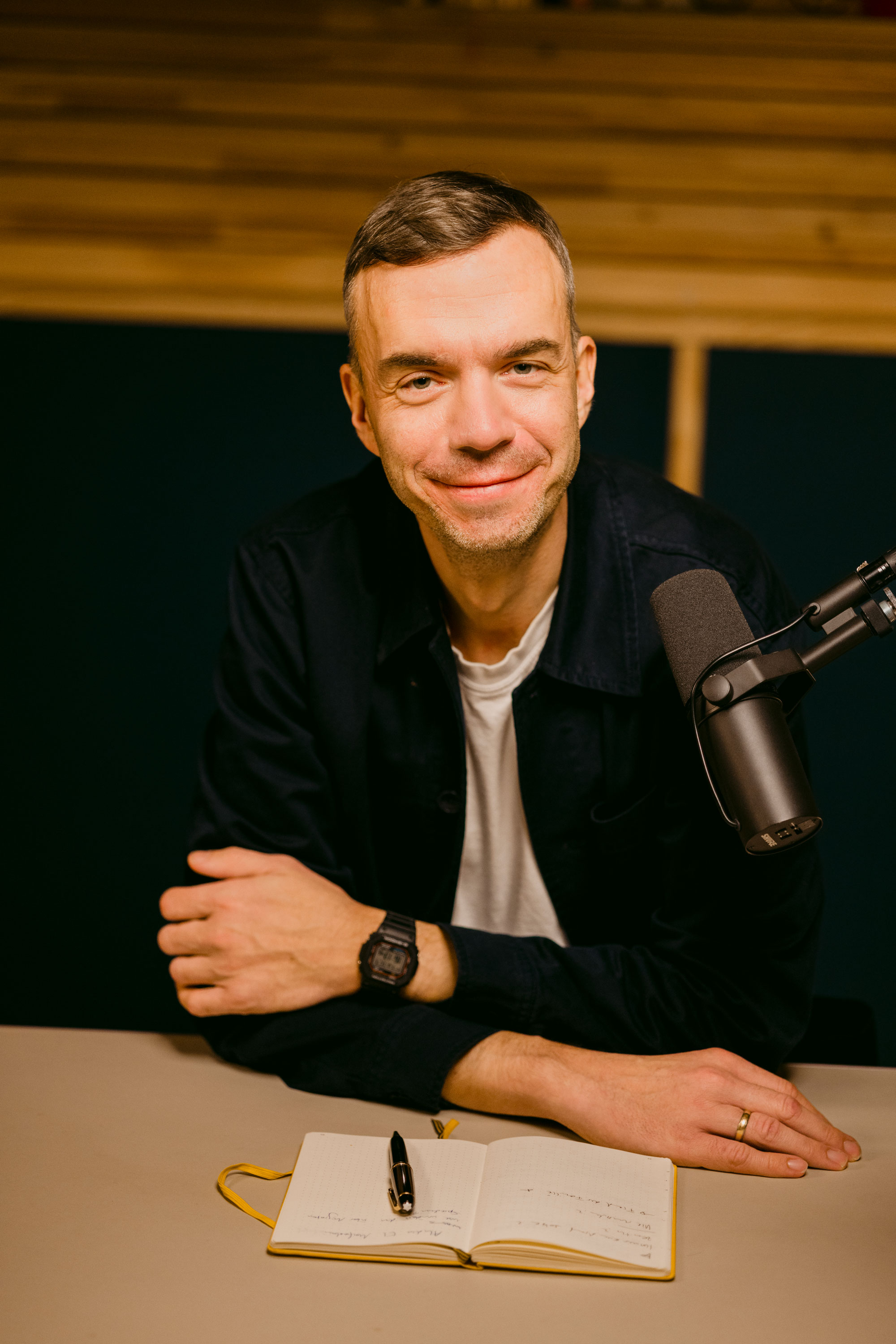 Person smiling, sitting at a table with a microphone and notebook.