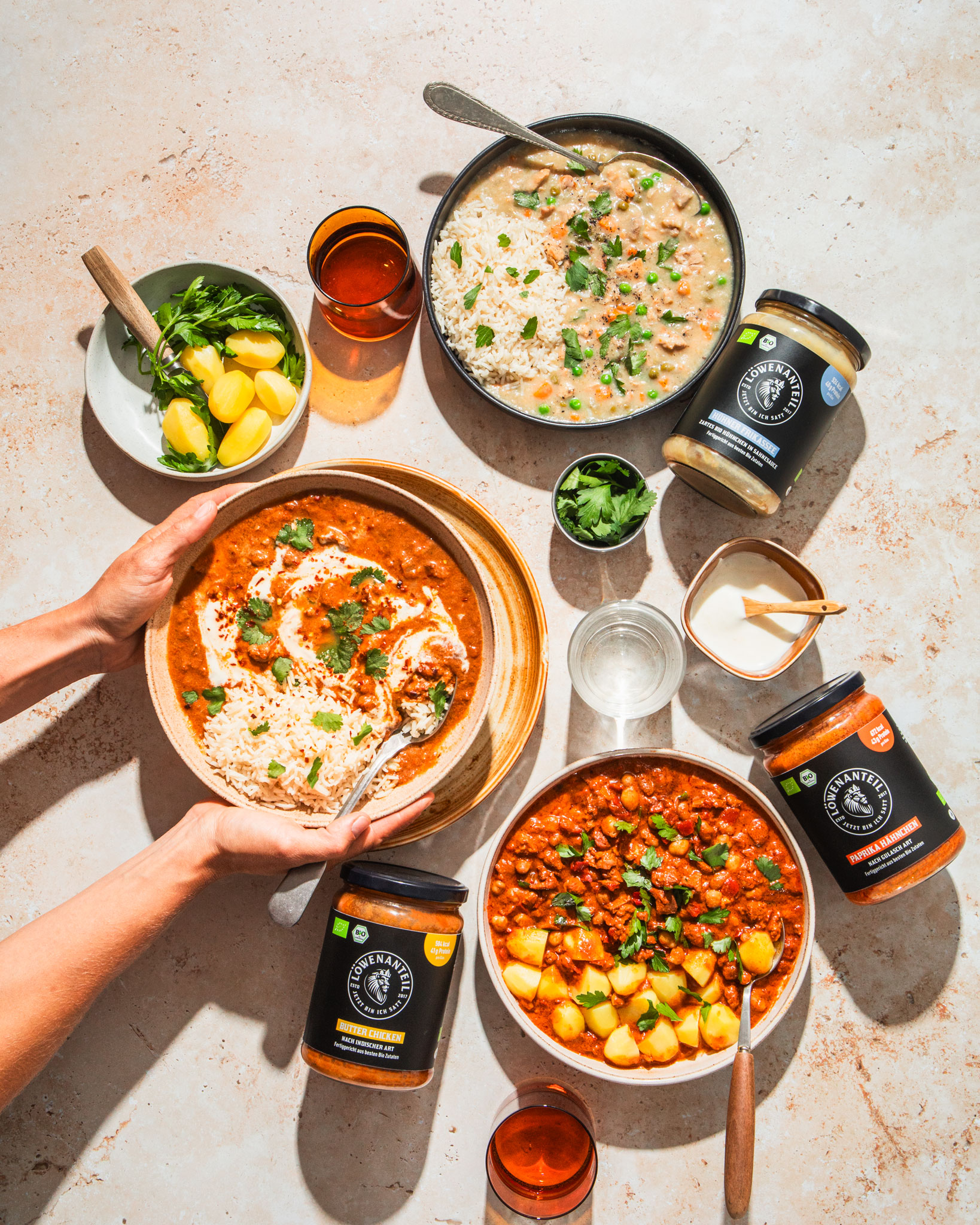 Dishes of curry and rice with garnish, surrounded by jars and drinks, hands serving a dish.