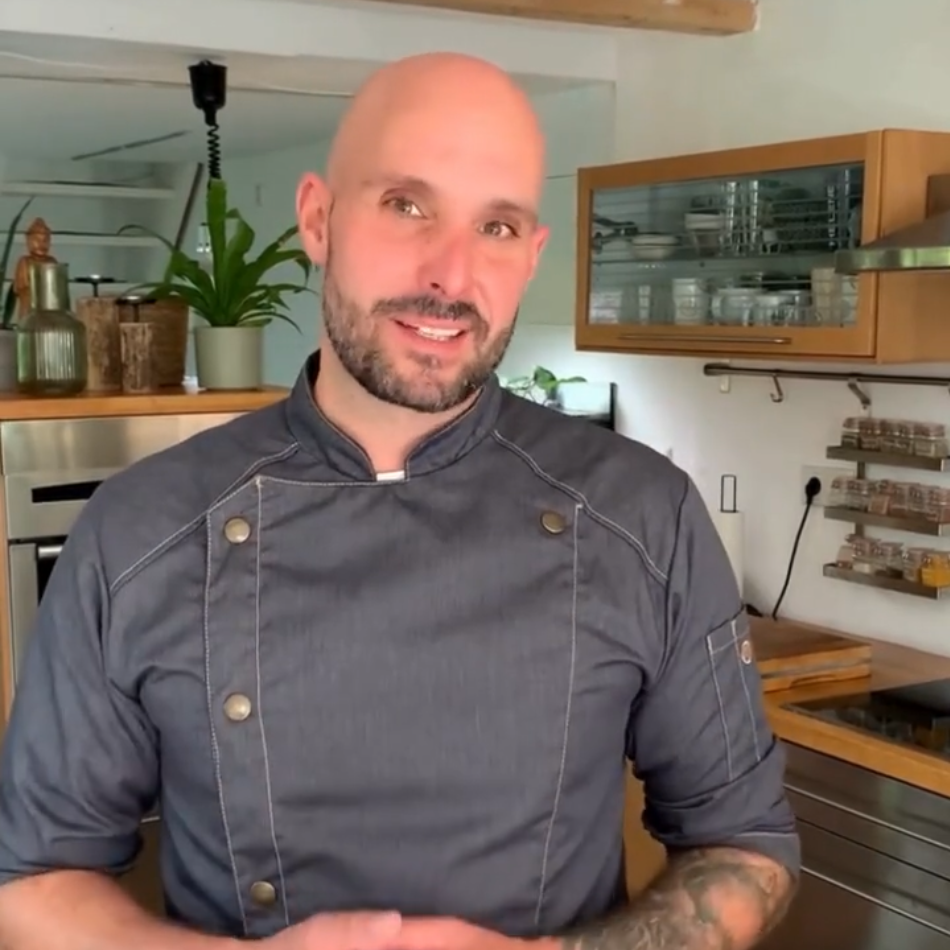 A bald man with a short beard wearing a grey chef's coat stands in a kitchen.
