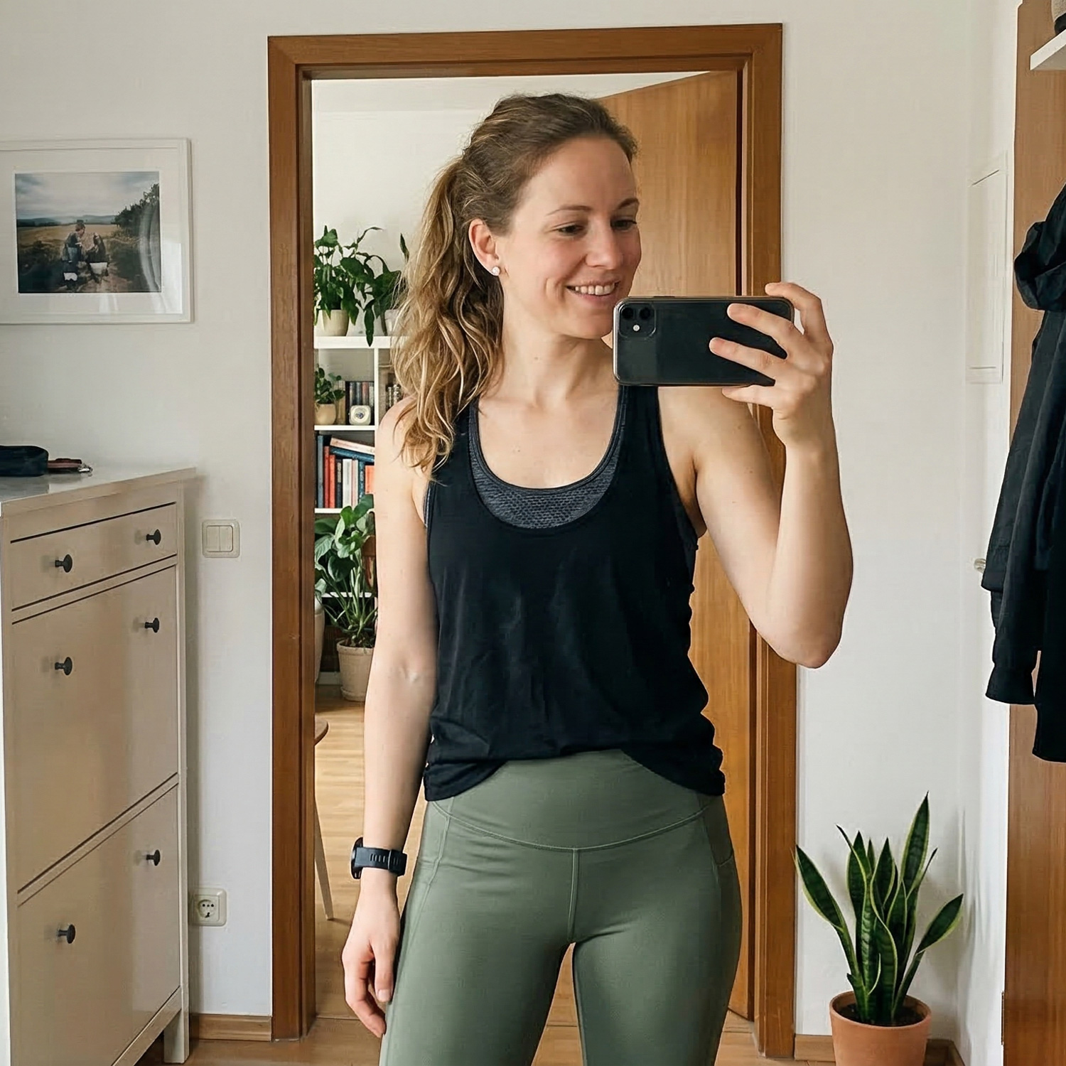 A woman in workout clothes smiles while taking a selfie in a mirror at home.