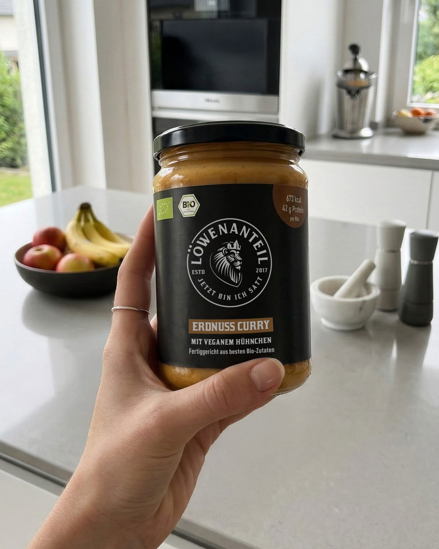 A person's hand holding a jar of Löwenanteil brand Erdnuss Curry in a kitchen.