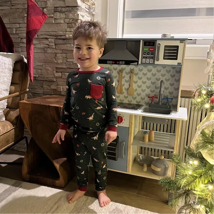 A young boy in dinosaur pajamas stands smiling next to a new toy kitchen.