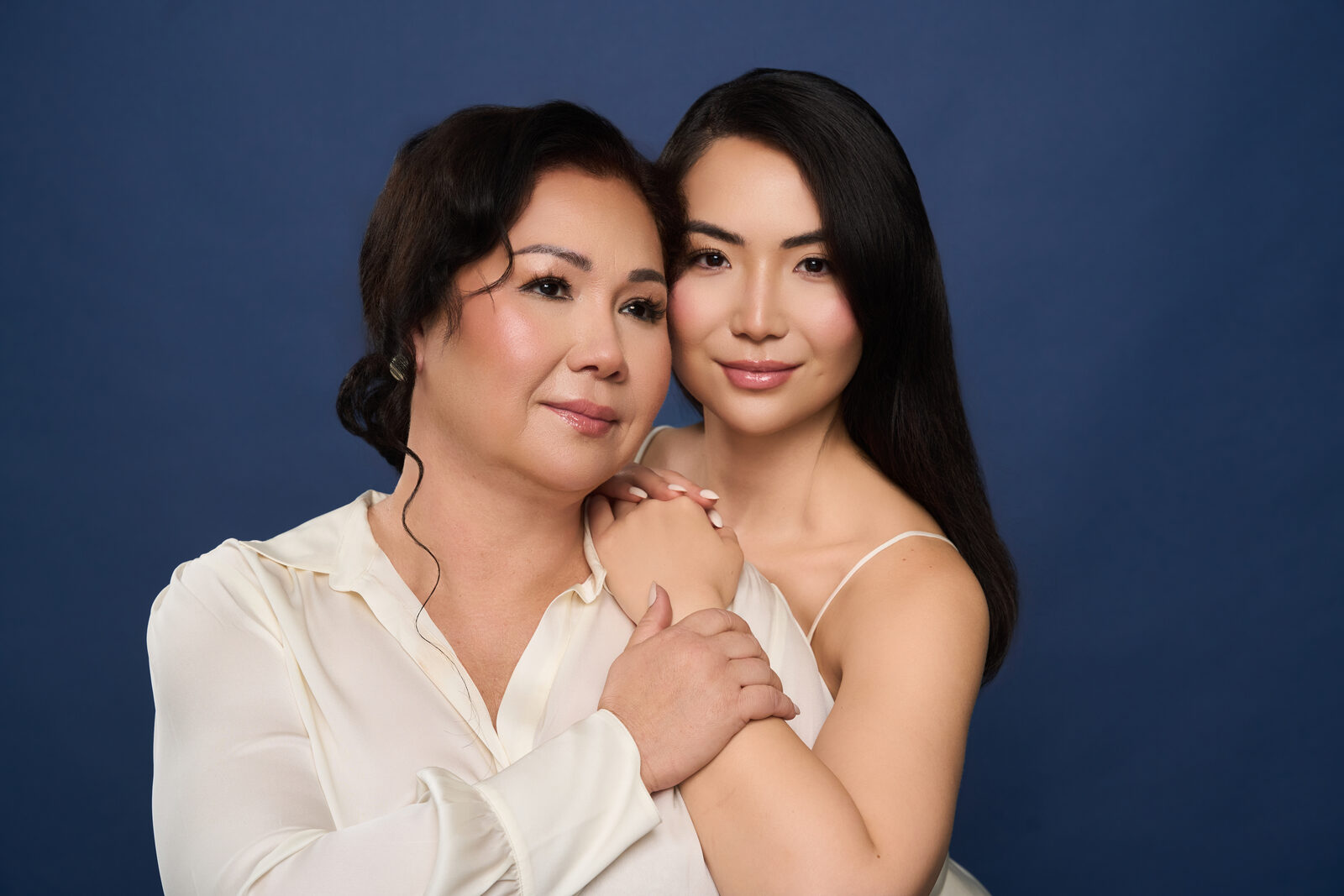 A young Asian woman embraces an older Asian woman from behind against a solid blue background.