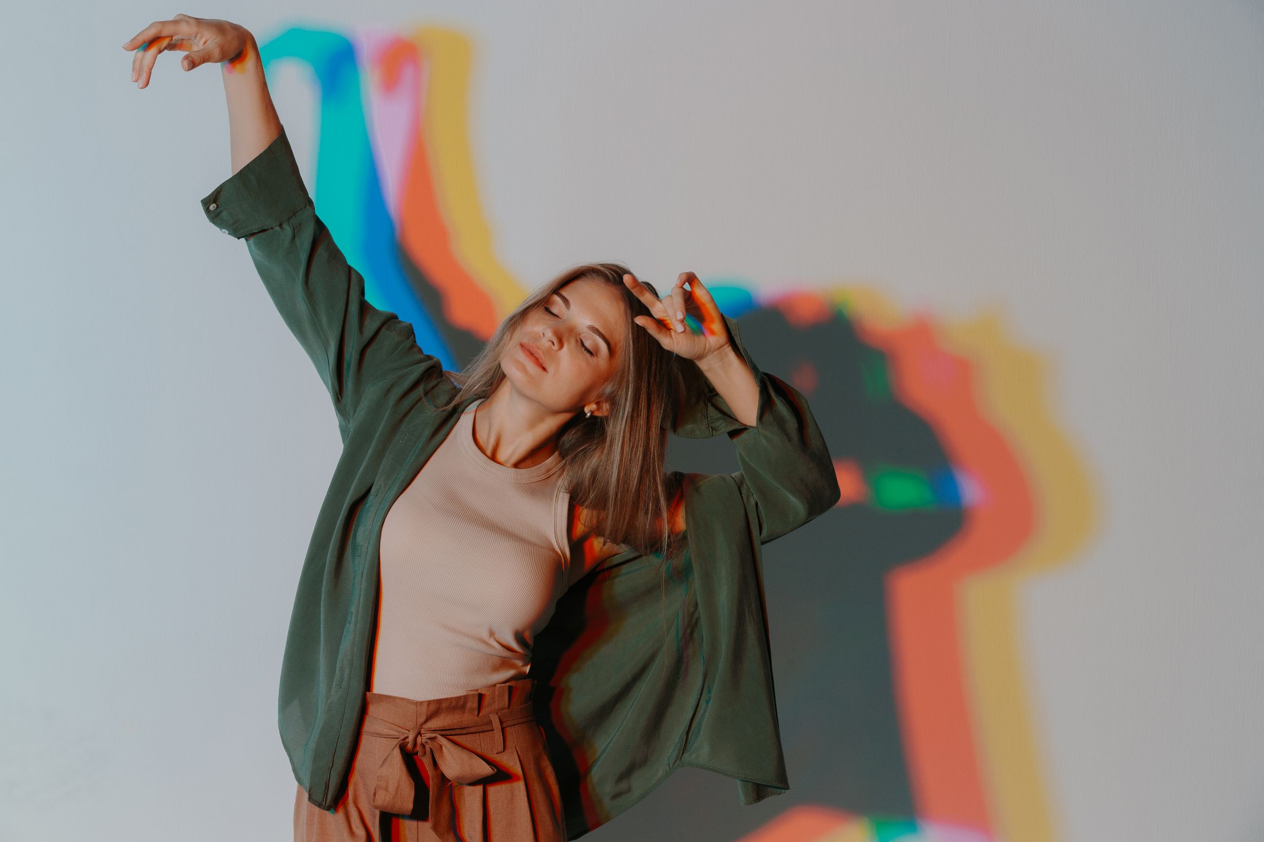 A woman with her eyes closed poses with her arms up against a wall with a colorful light projection.