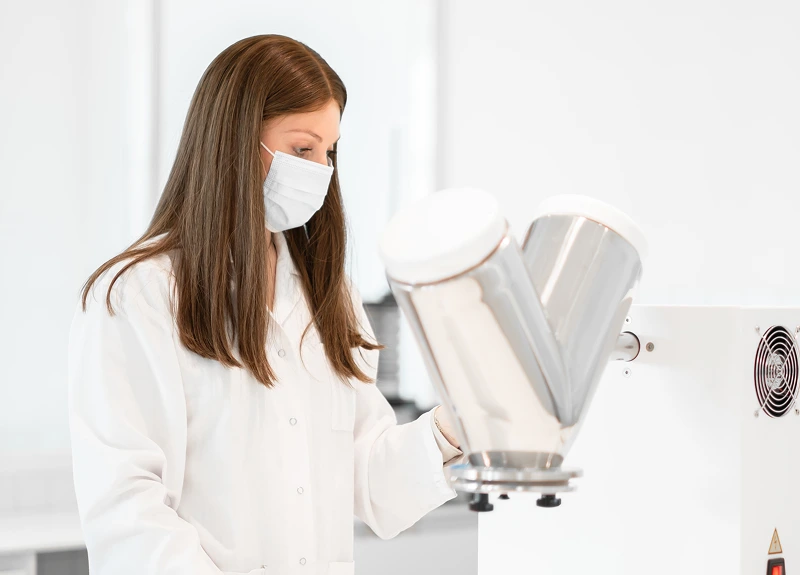 Person in laboratory coat and mask examining scientific equipment.