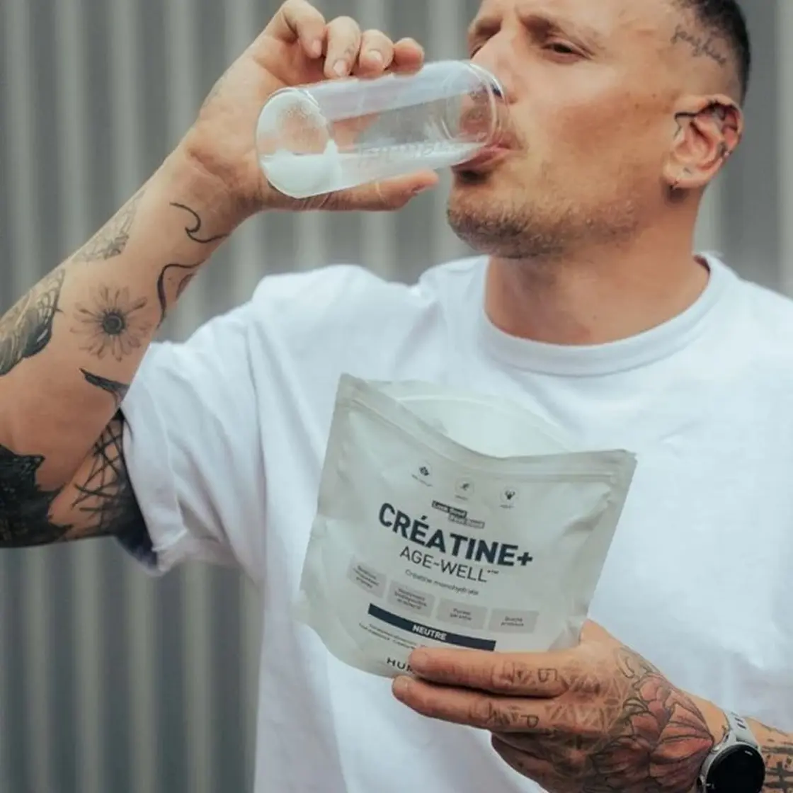 A tattooed man drinks a supplement from a glass while holding a white pouch of Créatine+ Age-Well.