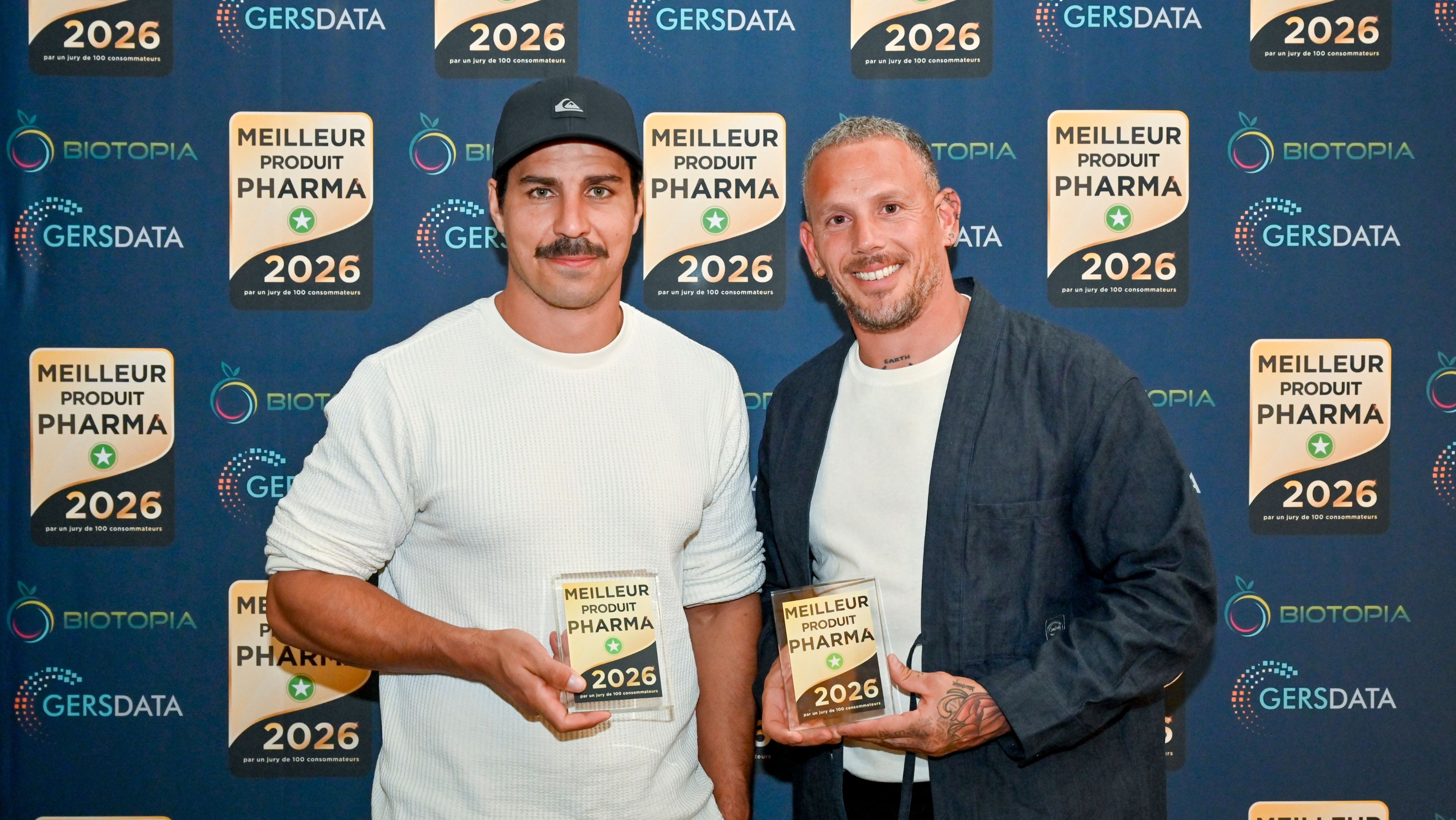 Two men holding awards, standing in front of a promotional backdrop.