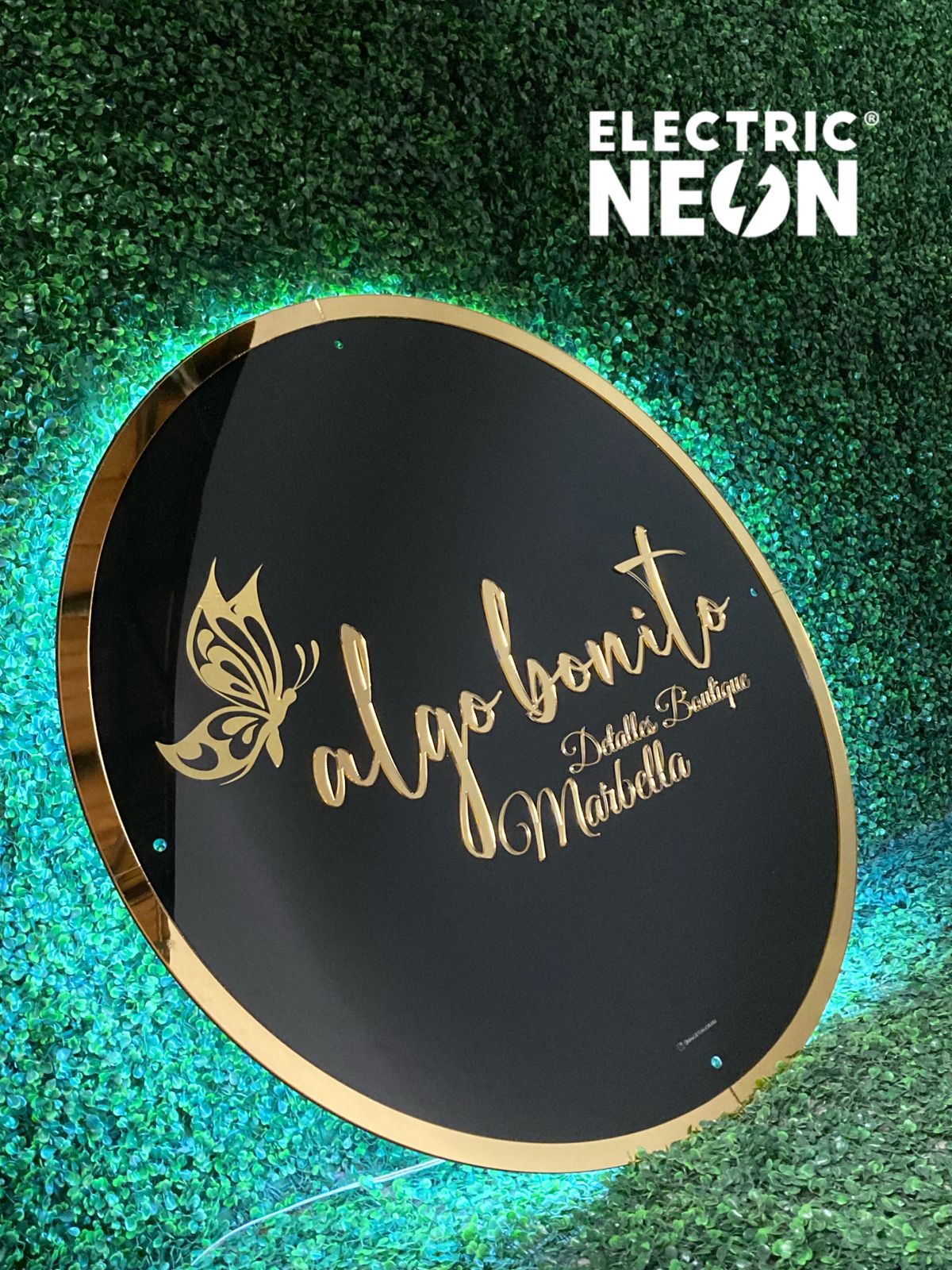 Round black sign with gold text and butterfly, on a green leafy background.