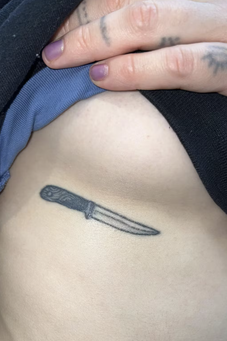 A close-up of a small black and grey knife tattoo on a person's side under their breast.